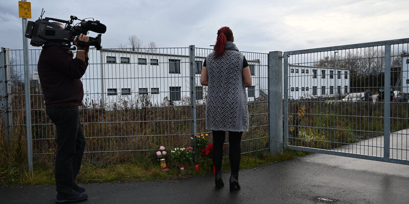 Ein Kameramann und eine Frau sind von hinten zu sehen, sie stehen am Zaun vor einer Unterkunft für Wohnungslose