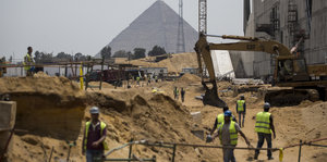 Bauarbeiter auf einer Baustelle im Sand, dahinter: die Pyramiden von Gizeh.
