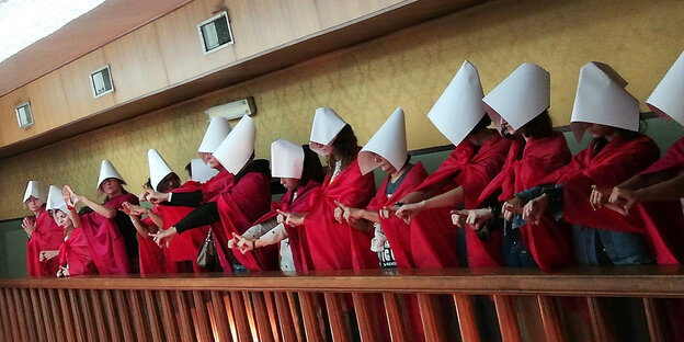 Frauen der italienischen feministischen Gruppe „Non una di meno“ in roten Roben und mit weißen Spitzhauben stehen nebeneinander auf einer Brüstung