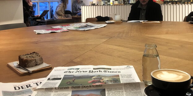 Blick auf einen Tisch, darauf eine Zeitung, Kaffeetasse, Stück Kuchen, im Hintergrund Cafégäste