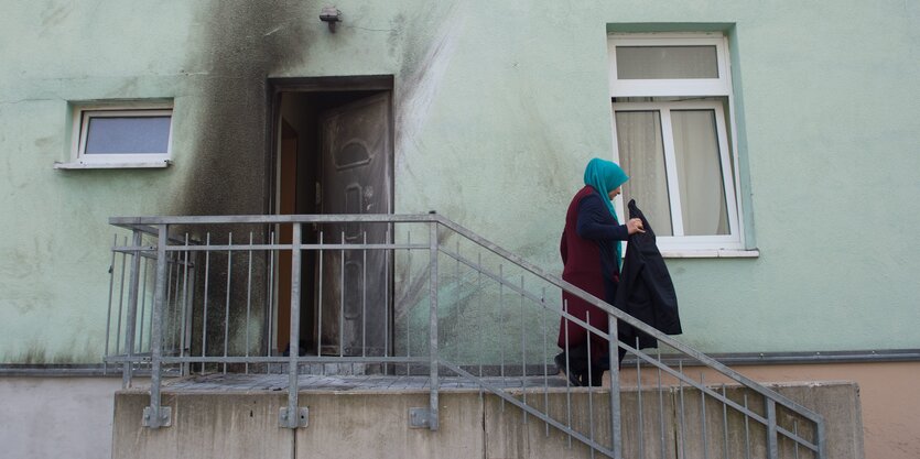 Eine Frau mit Kopftuch geht eine Treppe hinunter, an dem Gebäude sind Spuren eines Brandanschlags zu sehen