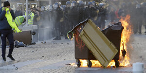 Im Vordergrund eine brennende Mülltonne und ein Protestler, der sich die gelbe Weste vor die Nase hält. Im Hintergrund Polizei