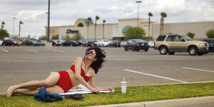 Eine Frau liegt im Bikini auf einem Grünstreifen auf dem Parkplatz eines Einkaufszentrums