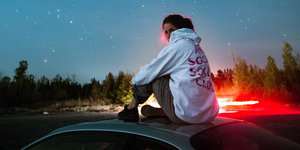 Eine junge Frau sitzt auf einem Autodach vor einem Sternenhimmel