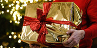 Ein Mann hält ein golden eingepacktes Weihnachtsgeschenk in den Händen. Im Hintergrund ist ein Weihnachtsbaum zu sehen.