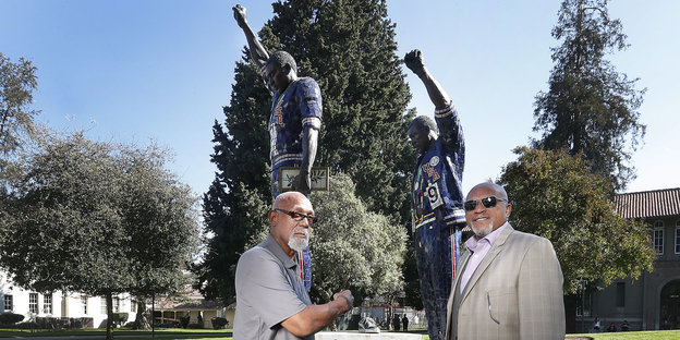 Tommie Smith und John Carlos stehen vor dem Denkmal, der sie in ihrer Pose mit ausgestreckter Faust bei der Siegerehrung zeigt