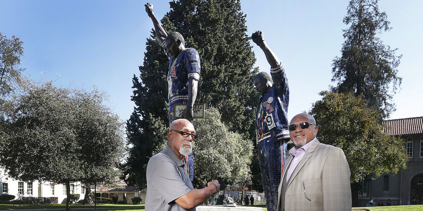 Tommie Smith und John Carlos stehen vor dem Denkmal, der sie in ihrer Pose mit ausgestreckter Faust bei der Siegerehrung zeigt