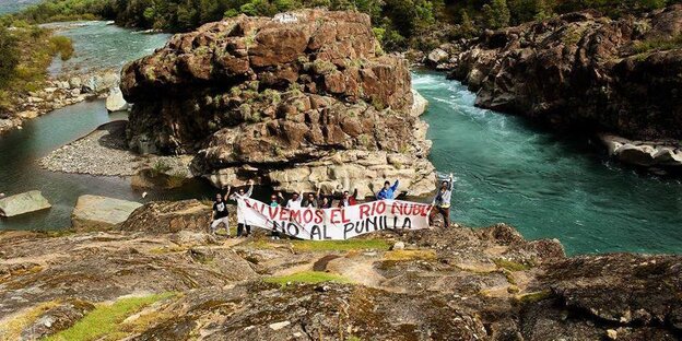 Eine Protestgruppe steht vor einem Fluß und hält ein Banner hoch