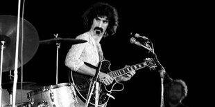 Frank Zappa steht vor einem Mikrofon und hat eine Gitarre in der Hand