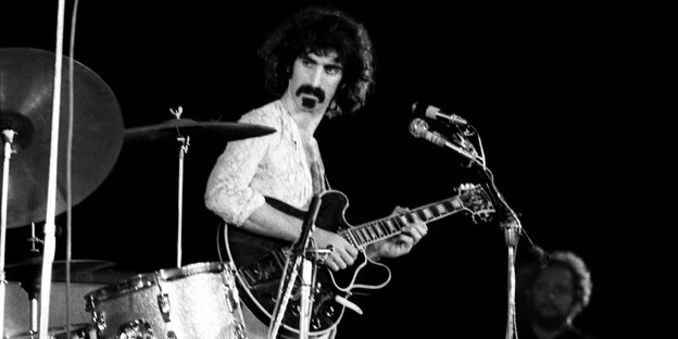 Frank Zappa steht vor einem Mikrofon und hat eine Gitarre in der Hand