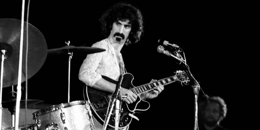 Frank Zappa steht vor einem Mikrofon und hat eine Gitarre in der Hand