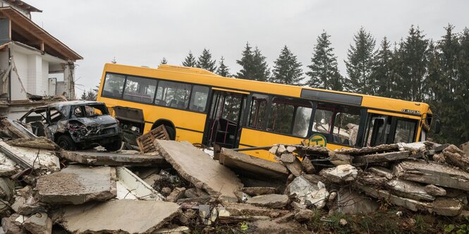 Neben einem zerstörten Gebäude stehen ein kaputter Bus und ein kaputtes Auto