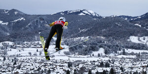 Mann auf Skiern fliegt ins Tal. Im Hintergrund: Berge