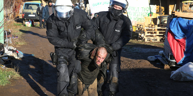 Zwei Polizisten mit Helm halten zwischen sich einen Mann fest, der kniet