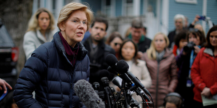 Elizabeth Warren spricht in mehrere Mikrofone, hinter ihr stehen viele Menschen