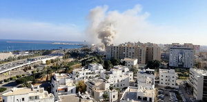 Ansicht von oben auf Tripolis, links im Bild das Meer. Im Zentrum der Aufnahme steigt weißer Rauch auf. Es herrscht blauer Himmel