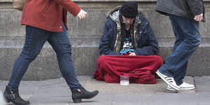 Mann hockt auf der Straße, teils verhüllt in roter Decke. Zwei Menschen eilen vorbei, man sieht nur ihren Unterleib