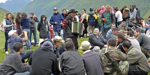 Ganz viele Leute sitzen und stehen im Freien in einer bergigen Region. Ein älterer Mann mitten unter ihnen spielt Akkordeon