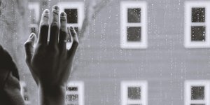 Eine Schwazweißbild zeigt eine Frauenhand, die an einer Fensterscheibe mit Regentropfen lehnt