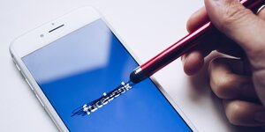 Ein weißes Smartphone, auf dem Bildschirm ist der Schriftzug Facebook zu lesen, der von einem Stift durchgestrichen wird.