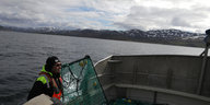 Ein Mann steht mit Netzen auf einem Fischerboot, das vor Bergen auf dem Meer treibt