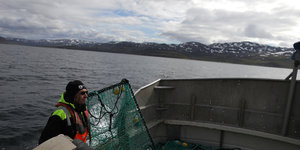 Ein Mann steht mit Netzen auf einem Fischerboot, das vor Bergen auf dem Meer treibt