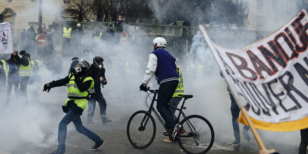 Demonstranten mit gelben Westen stehen in einer Tränengaswolke, eine Person hält ein Transparent hoch