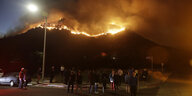 Menschen stehen bei Nacht vor einem Hügel, auf dem Wald brennt