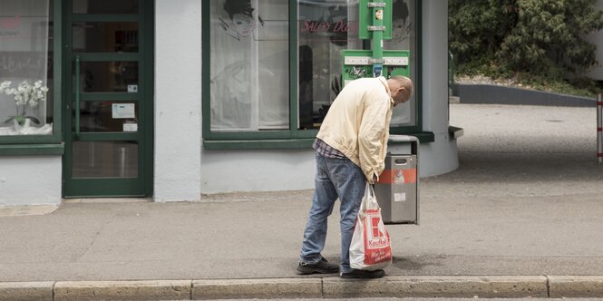 Ein Mann hat eine Plastiktüte in der Hand und hält seine Hand in einen Mülleimer