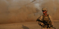 Ein US-amerikanischer Soldat steht in der Wüste, im Hintergrund wirbelt ein Helikopter Staub auf