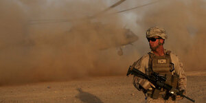 Ein US-amerikanischer Soldat steht in der Wüste, im Hintergrund wirbelt ein Helikopter Staub auf