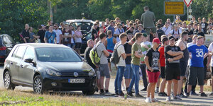 Eine große Gruppe junger Menschen steht auf einer Straße neben einem Auto und einem Ortsschild
