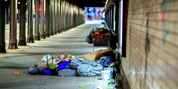 Ein Obdachloser liegt mit Schlafsack, Decken und mehreren Flaschen unter einer Eisenbahnunterführung.