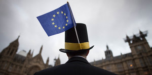 Ein Mann mit Hut und kleiner EU-Fahne steht vor dem britischen Parlament, er ist von hinten unten zu sehen