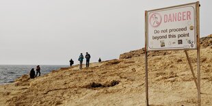 an einem Strand, mehrere Leute im Hintergrund, ein Schild mit Gefahrenwarnung im Vordergrund