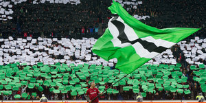 Mann schwenkt Fahne in Vereinsfarben schwarz-weiß-grün vor Fanblock von Hannover 96