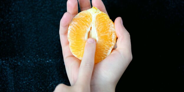 Eine halbe geschälte Orange in der einen Hand, zeigt der Zeigefinger der anderen Hand auf deren Mitte