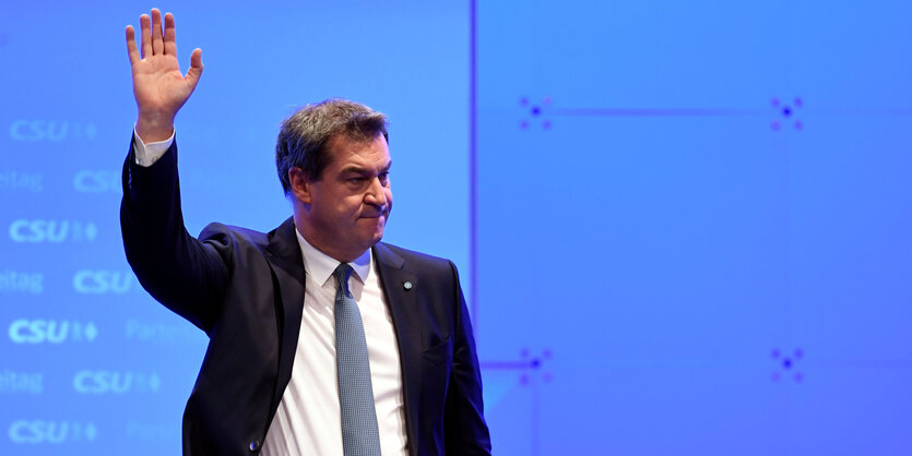 Markus Söder winkt vor blauem Hintergrund