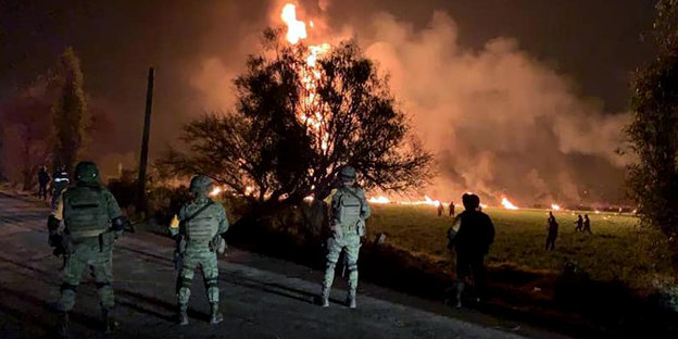 Soldaten stehen auf einer Straße in einigem Abstand zu meterhohen Flammen