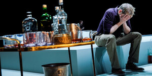 Ein Mann sitzt neben einem Haufen leerer und voller Flaschen auf der Bühne und vergräbt sein Gesicht in den Händen