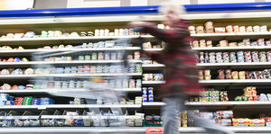 Eine Frau mit Enkaufswagen huscht in einem Supermarkt an einem Regal mit Waren vorbei.