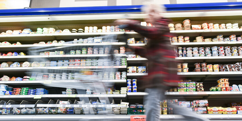 Eine Frau mit Enkaufswagen huscht in einem Supermarkt an einem Regal mit Waren vorbei.