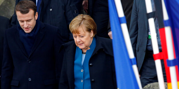 Merkel und Macron stehen in schwarzen Jacken am linken Bildrand, rechts davon Flaggen