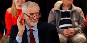Jeremy Corbyn kneift die Augen zusammen und hält die Hand am Ohr. Hinter ihm sitzen Menschen