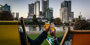 Zwei Männer im Liegestuhl prosten sich zu, im Hintergrund, der Main und die Kulisse der Frankfurter Innenstadt