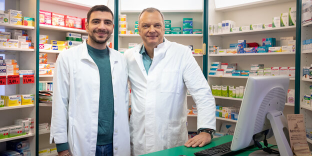 Zwei Apotheker stehen in Kitteln vor Regalen mit Medikamenten