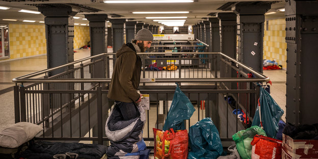 Ein Mann steht in einem U-Bahnhof zwischen Schlafsäcken und Tüten. Im Hintergrund sind Isomatten und weitere Schlafsäcke zu sehen