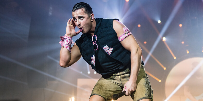 Andreas Gabalier trägt eine Lederhose und hält sich eine Hand an sein Ohr