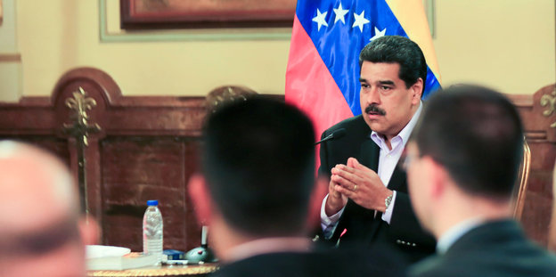 Nicolas Maduro sitzt an ein em Schreibtisch, mehrere Menshcen hören ihm zu