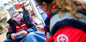 Eine Helferin spricht mit einem Mann, der draußen auf dem Boden vor einem Geschäft sitzt